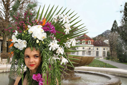 Florale Pracht auf dem Kopf platziert. Model Jessika vor dem Alpensolebrunnen des Königlichen Kurgartens Bad Reichenhall, dahinter die Konzertrotunde © Fischer/alpgold